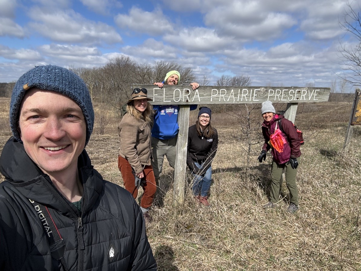 Photo of Prairie Pasqueflower monitoring team smiling in front of the Flora Prairie Preserve sign. Left to right: Grant Fessler, Jessie Crow-Mermel, Daniel Pohl, Ingrid Felsl, and Gretel Kiefer.